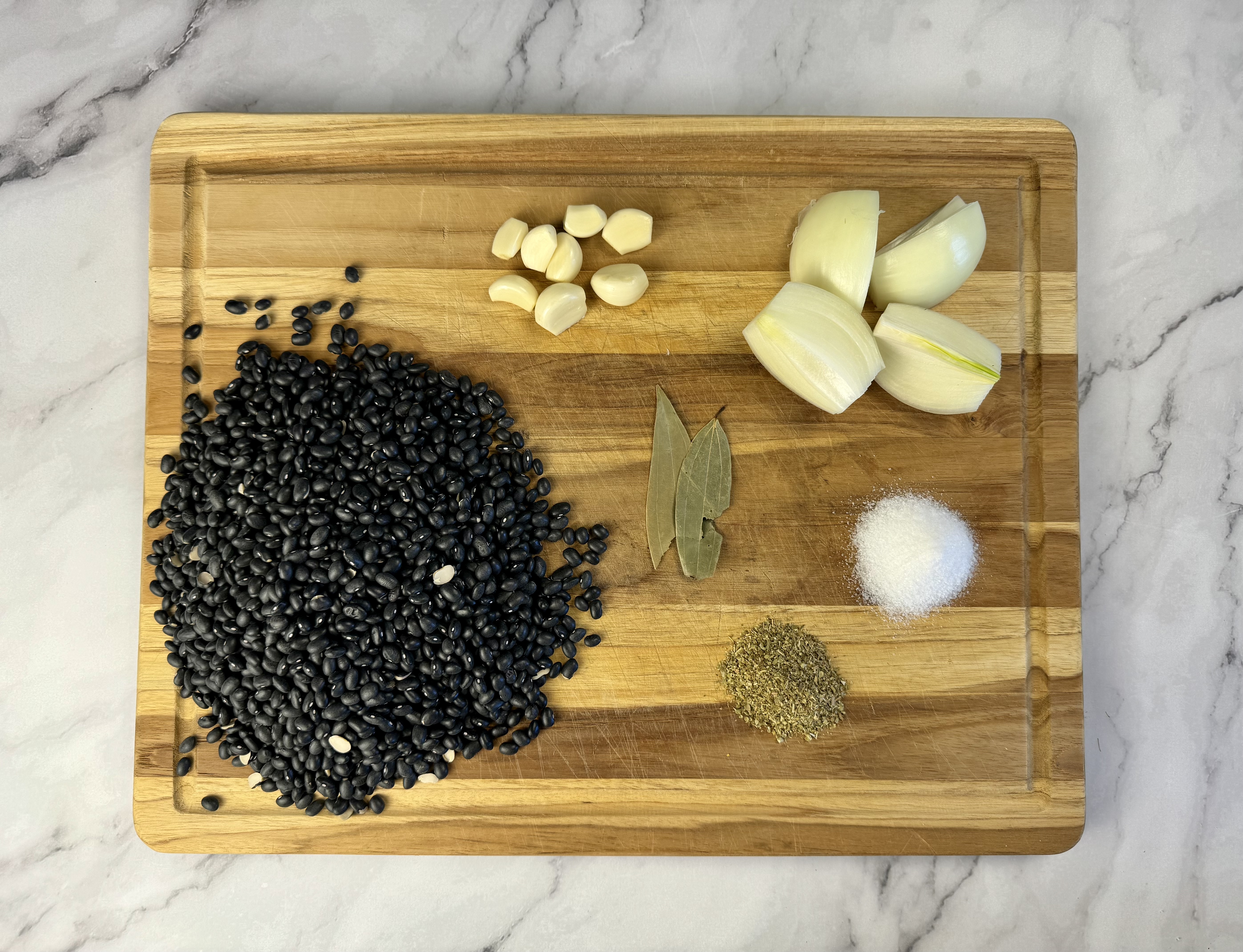 Ingredients for slow cooker black beans. Beans, garlic cloves, quartered onion, bay leaves, salt and oregano are on a cutting board on the kitchen counter.