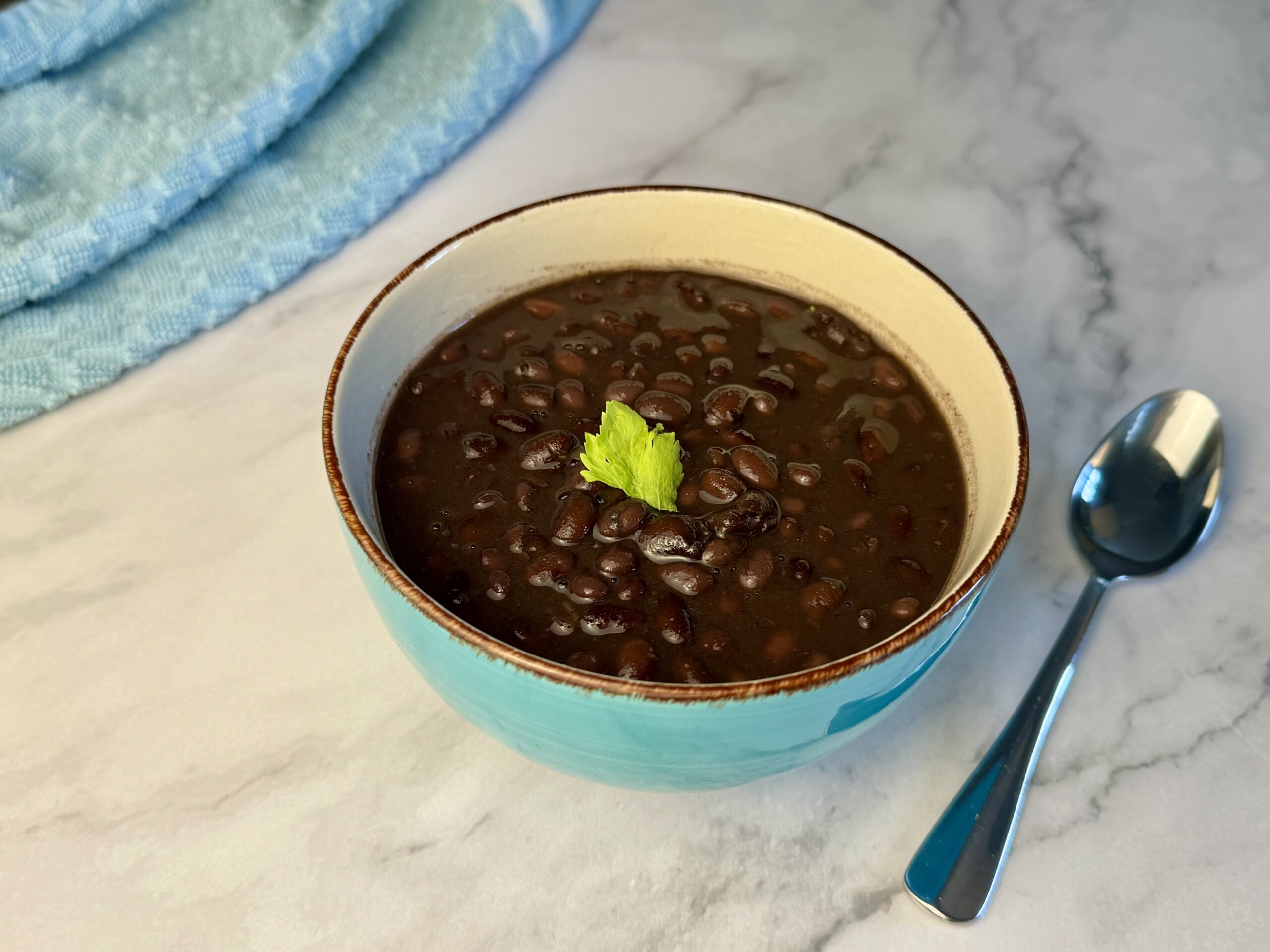 Black Beans with celery leaf garnish in a vibrant blue bowl on white marbled stone counter top. Spoon and dish towel elegantly placed alongside in the background.